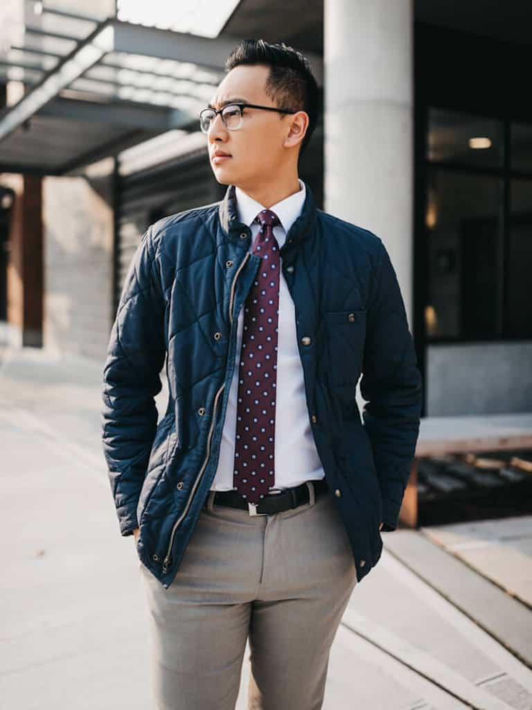 Person wearing a quilted jacket and tie standing on a sidewalk.