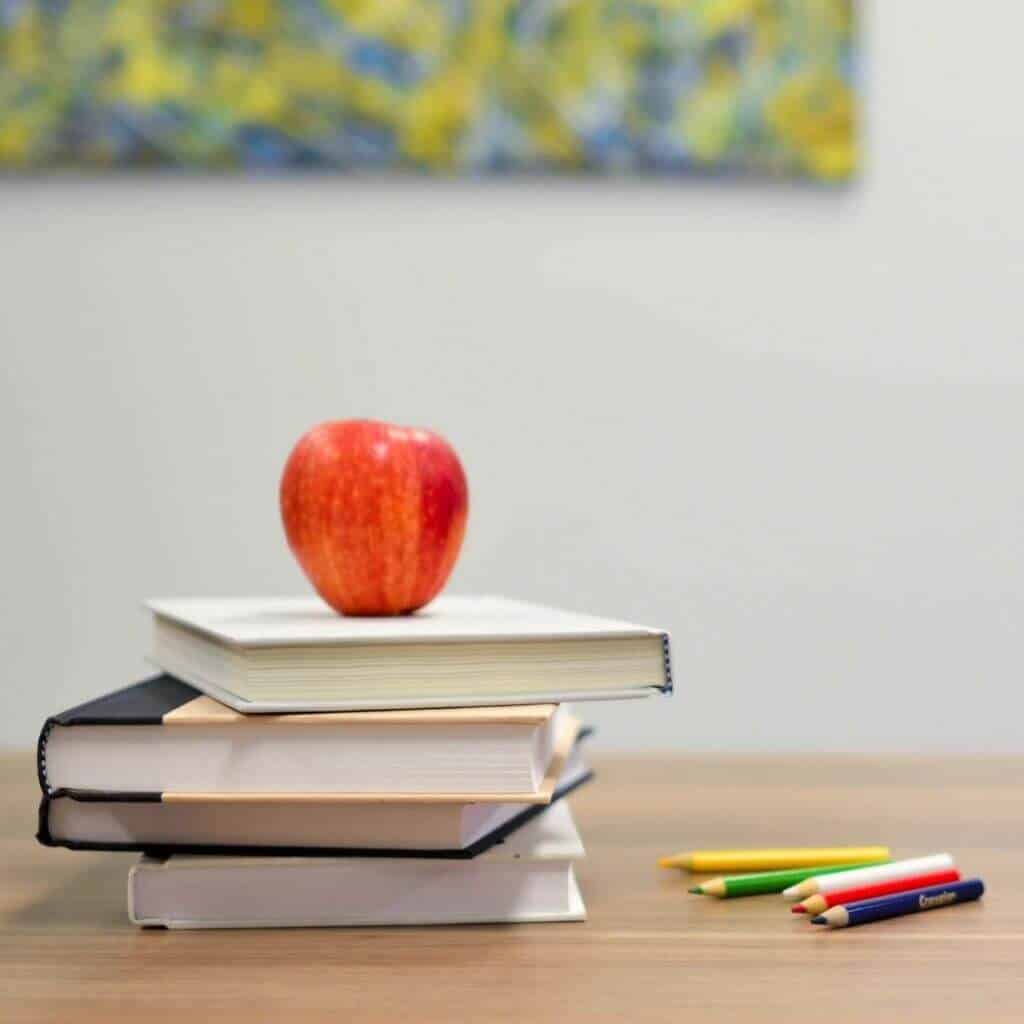 An apple on a stack of four books with colored pencils next to it.