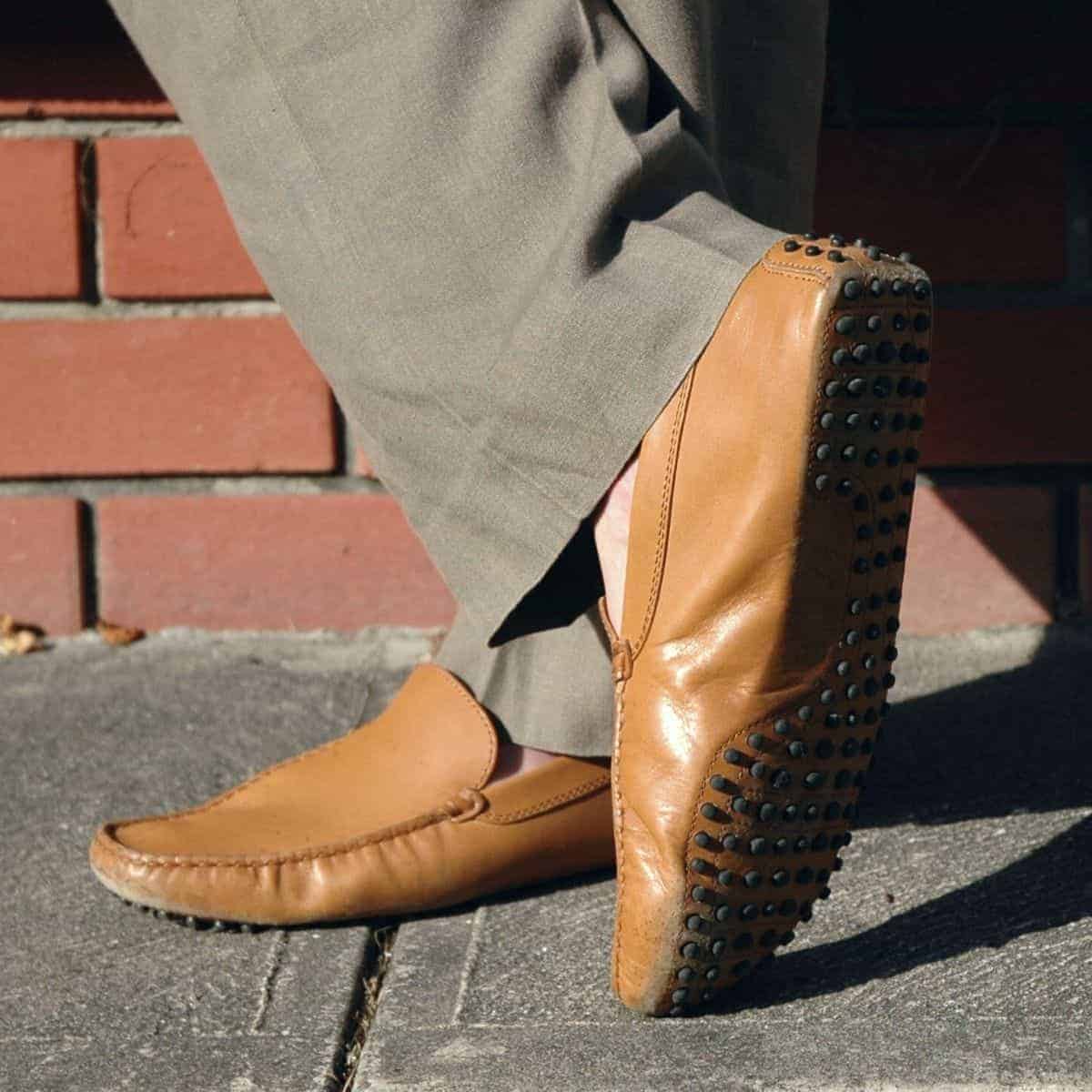 Close-up of a person wearing driving shoes with their legs crossed on a sidewalk.