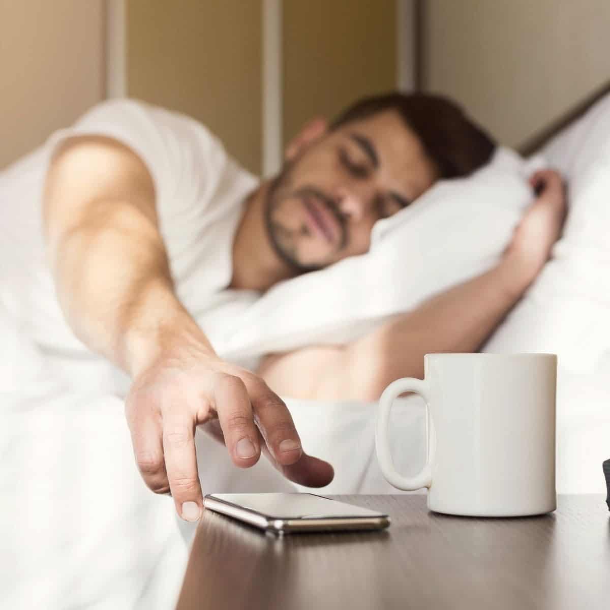 Person laying in bed and grabbing their phone from a table.